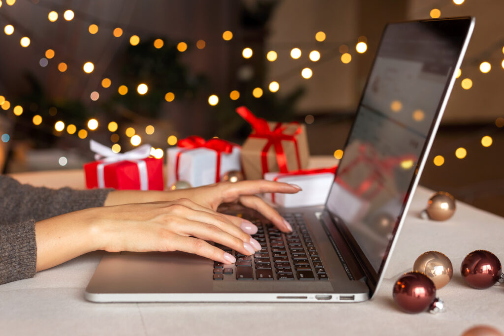 Hands are shown typing on a laptop with christmas lights and red wrapped gifts blurred in the background.