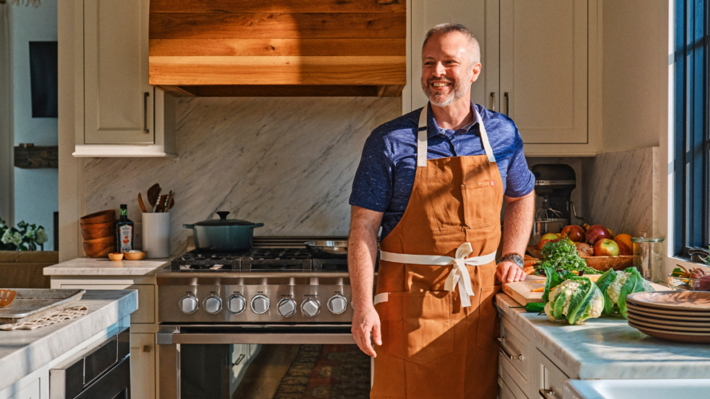 A middle aged man smiles and poses for a photo in a kitchen. He wears a dark orange apron.