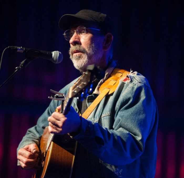 A man in denim jacket plays a guitar.