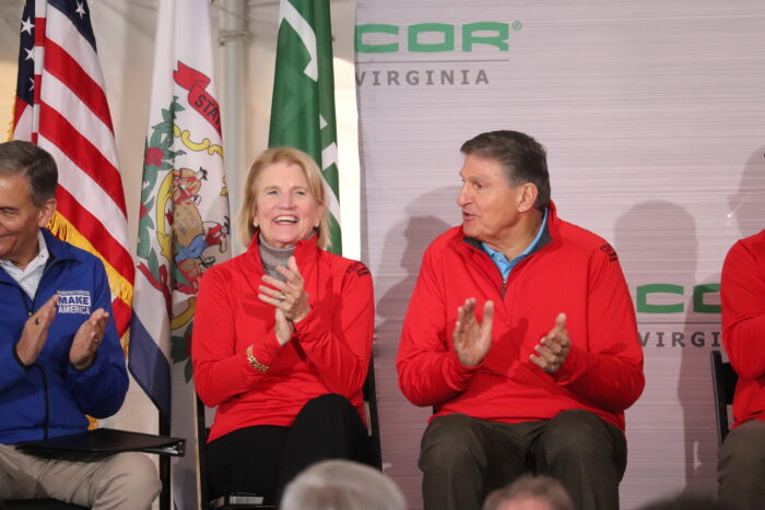 A man and a woman wearing red jackets are seated on a stage and applauding.