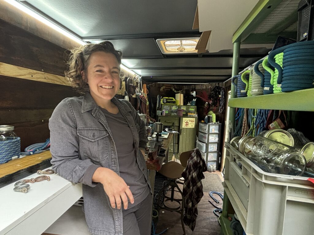 A woman leans against a shelf in a workshop filled with rock climbing gear.