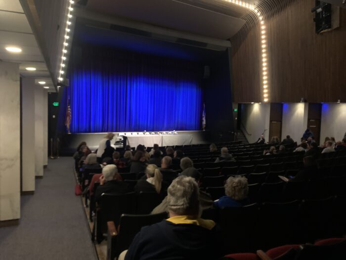 half-full auditorium, empty chairs behind a table on the stage, blue curtain in the back