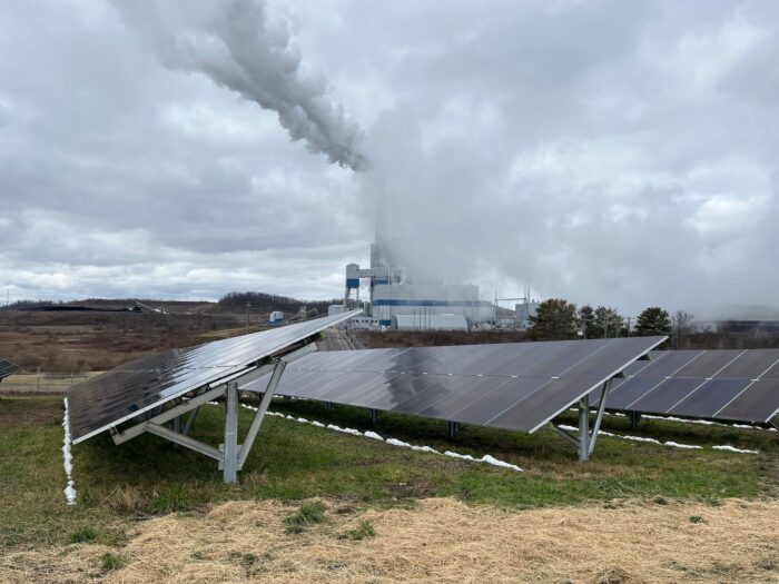 Steam rises above a power plant on an overcast day with an array of solar panels in the foreground.