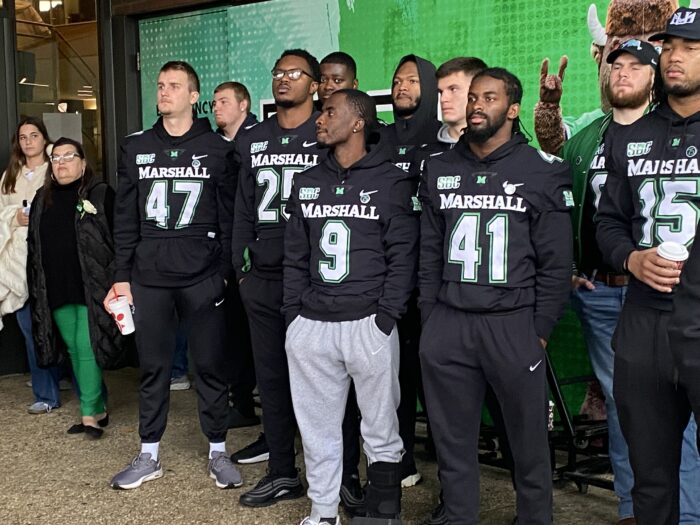 A group of young men wearing football sweatshirts stand together. The sweatshirts are black with white numbers and letters.