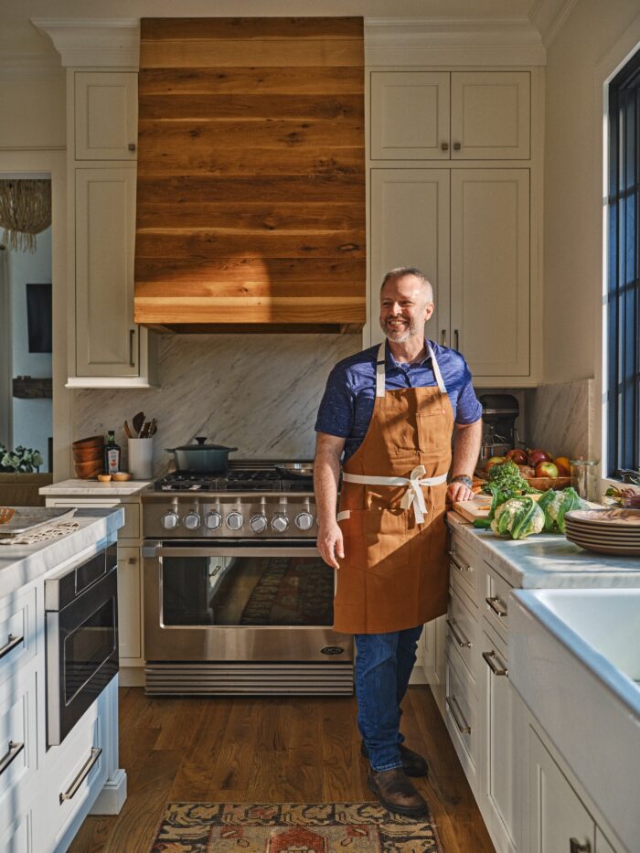 A man in blue with a brown apron stands by a kitchen counter covered with vegetables.