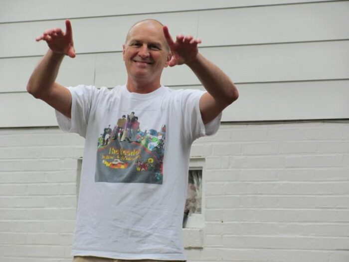 A smiling man in a white Beatles t-shirt holds his arm up as if directing an orchestra.