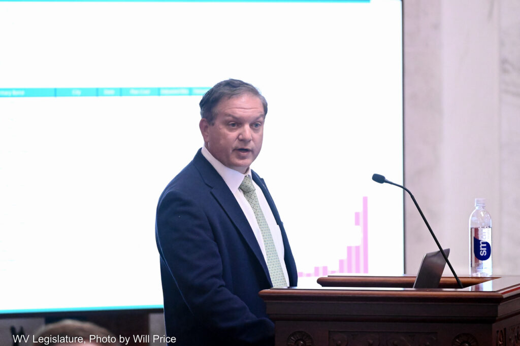 man speaks at a podium in front of a projector screen with graphs