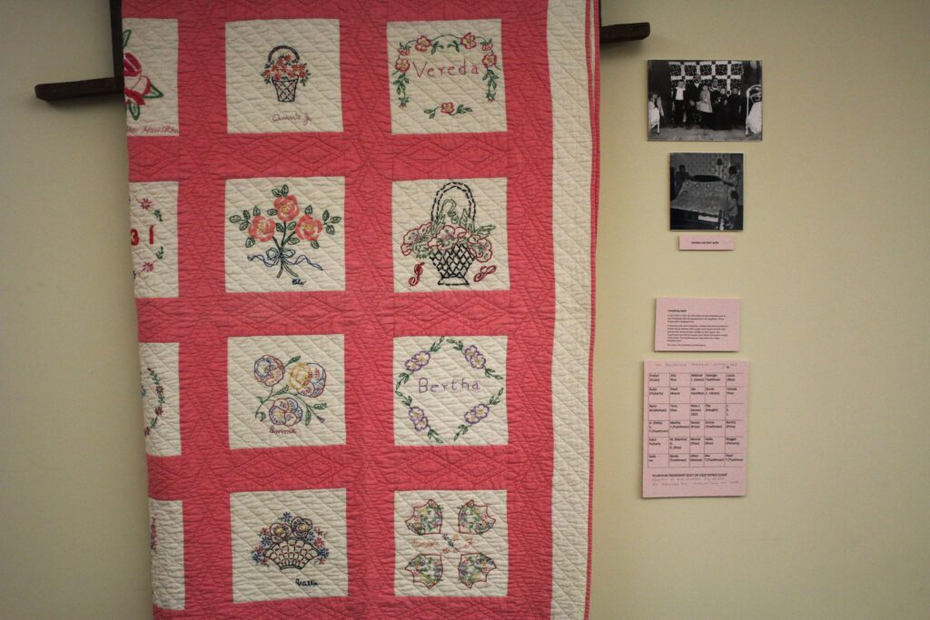 A pink and white border frames squares of floral embroidery on a quilt hanging on a wall. To the side of the quilt are photographs of the quilt's makers, as well as a map of the quilt showing who made what square.