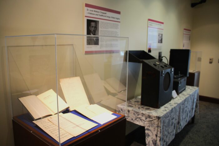 A display case holds musical papers. To the right of the display case, arrayed on a table with a botanical blue and white tablecloth, are midcentury portable recording equipment the size of modern luggage. The black rectangles have inlaid speakers and stand below informative posters hung on a beige wall.