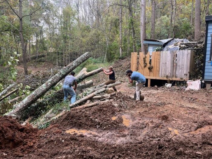 Two adults attempt to move fallen trees. One man is taking a photo of them.