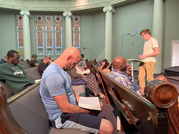 About a dozen men sit in a church listening to a speaker.