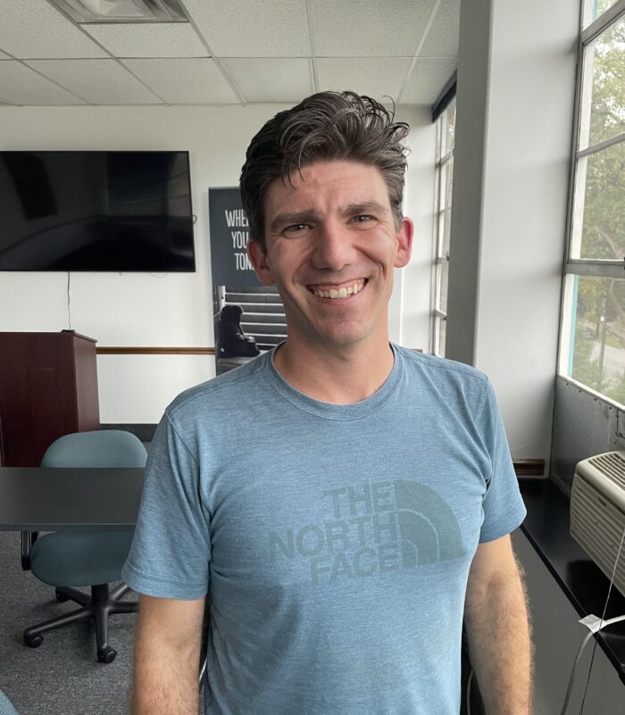 An adult man with brown hair smiles for the camera. He wears a blue t-shirt that says, "The North Face" on the front. Behind him is a conference room. Seen is a podium and a widescreen TV mounted on the wall.