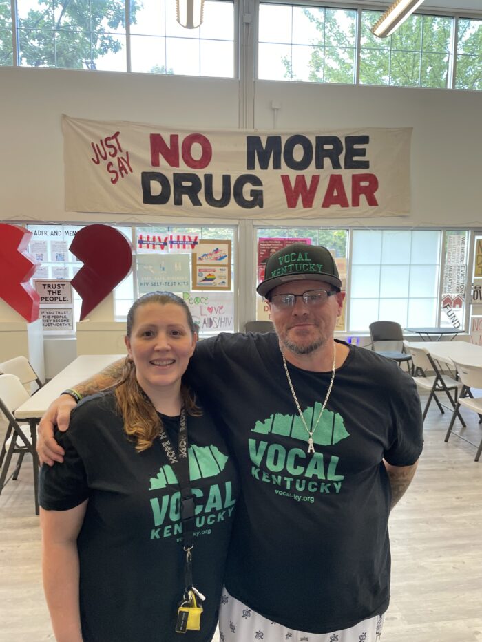 Two adults, a woman and a man, stand side by side and pose for a photo. Both wear t-shirts that say, "Vocal Kentucky." Behind them is a sign that reads, "Just Say No More Drug War."