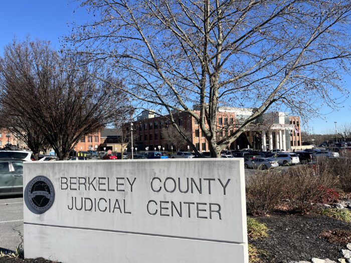 A concrete sign is engraved with the words "Berkeley County Judicial Center" and has a metal Berkeley County seal embedded to the left of the words. Behind it are a garden and a parking lot, and in the distance a red brick building with white columns and an American flag out front.