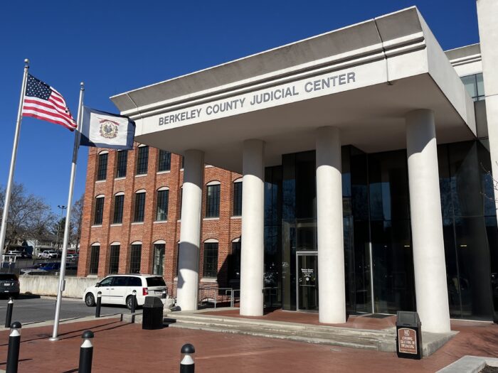 A red brick building has large white columns out front, with lettering above them that reads "Berkeley County Judicial Center." In front of the building is a red brick sidewalk and the American and West Virginia flags. Beneath the columns, large glass panes stretch across the length of the building.