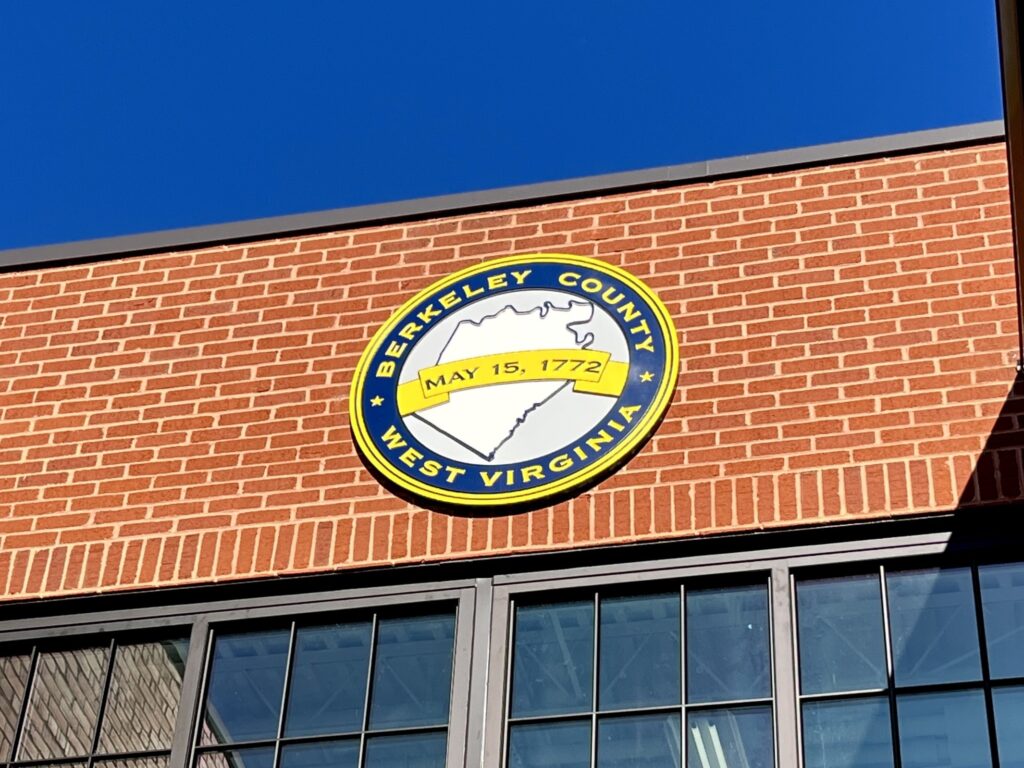 Atop a red brick building with black window panes, a blue and yellow metal seal shaped in a circle shows the outline of Berkeley County and reads "Berkeley County West Virginia." It also shows the date May 15, 1772, the date of the county's founding.