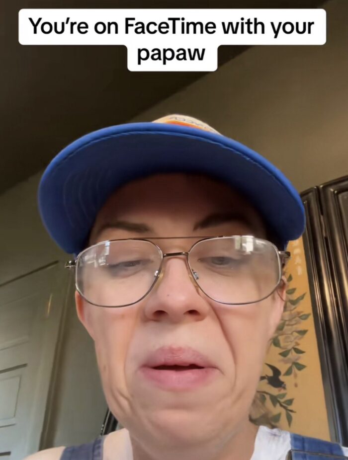 A closeup shot of an adult using a cell phone in a video call. The woman is playing a character and making a funny face. She wears glasses and a ball cap. Above her are the words, "You're on FaceTime with your papaw."