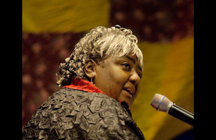 A woman sits in front of a microphone, in front of a purple and yellow quilt. She appears to be seated at a piano