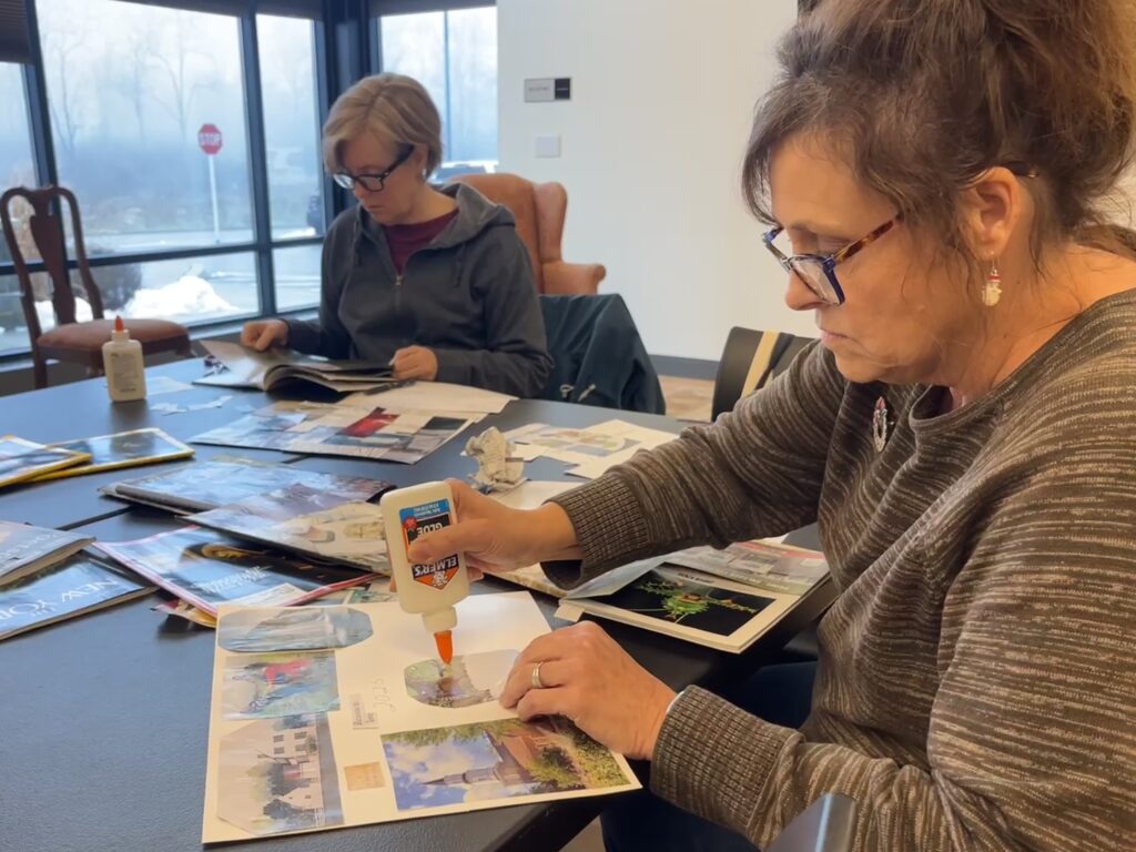 A woman with glasses and her hair in a bun glues a magazine cutout onto a white sheet of paper. She sits at a black table with many magazines placed on top of it. Another woman flips through a magazine on the other side of the table, in the background.