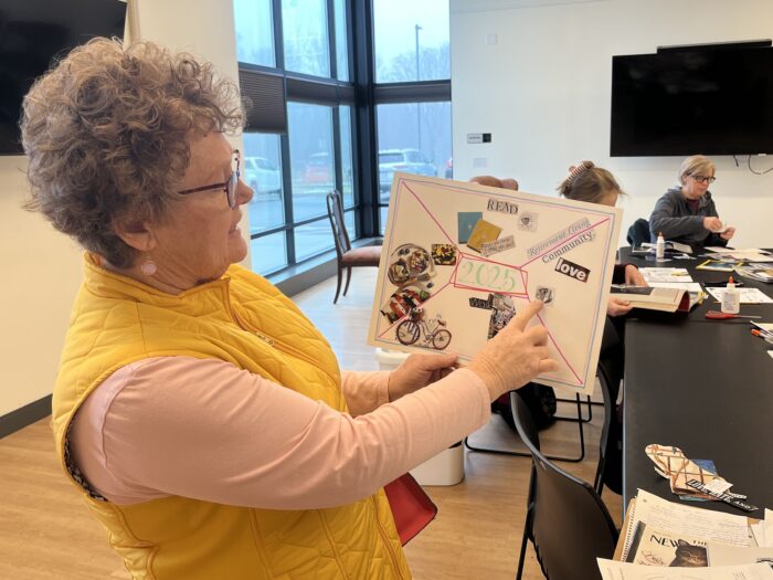 A woman in a yellow vest with short, gray hair holds up a piece of paper with magazine cutouts pasted onto it. She points at one image on the collage.