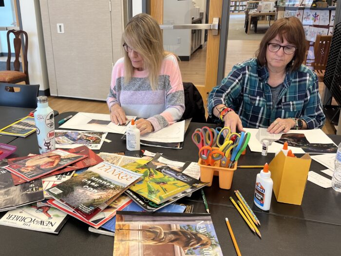 Two women sit at a black table with magazines, scissors and bottles of glue piled on top. Each has a white sheet of paper in front of them, and is gluing magazine clippings to the paper.