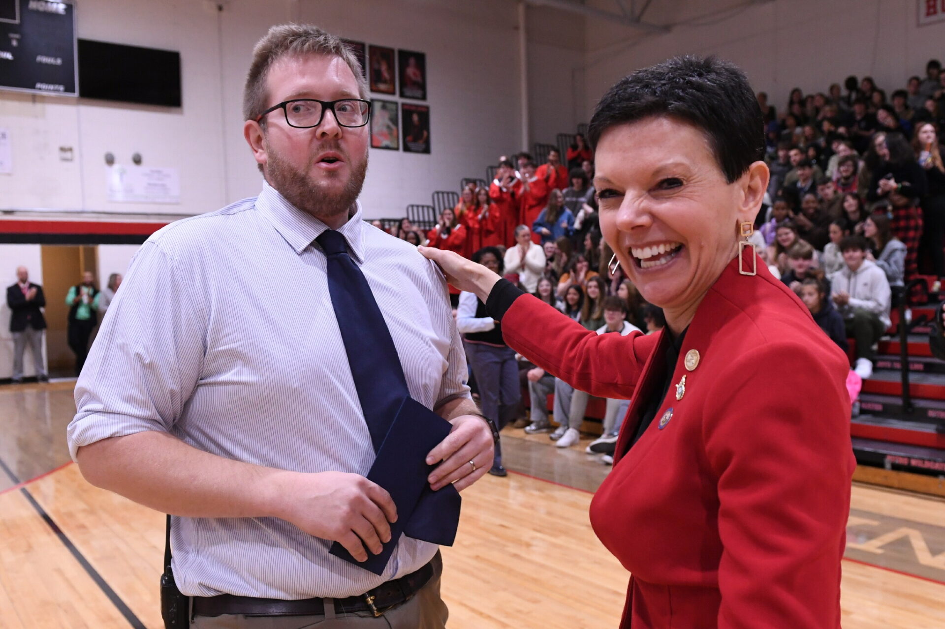 A woman wearing a red blazer with short black hair smiles broadly as she awards a man wearing a dress shirt and tie a $25,000 prize for excellence in teaching.