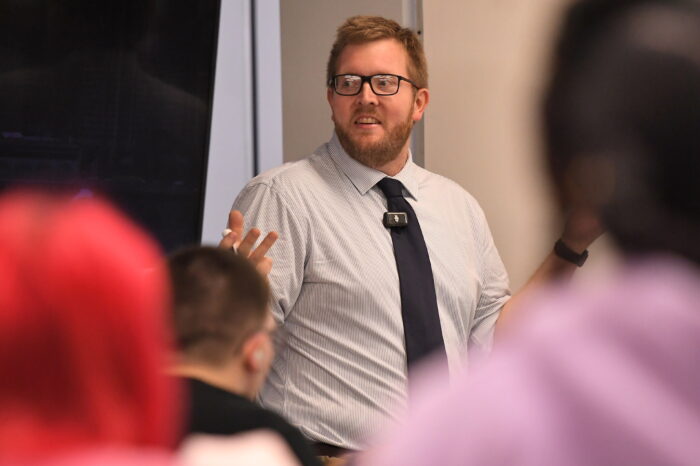 A blonde man with a beard wearing black-rimmed rectangle glasses speaks at the front of a classroom.