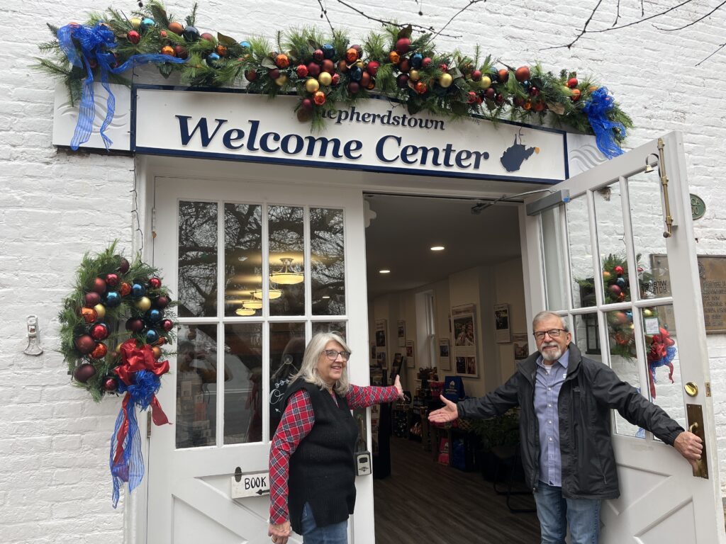 A woman and man stand outside a white building labeled "Shepherdstown Welcome Center." They stand on either side of the entryway doors, gesturing inside.