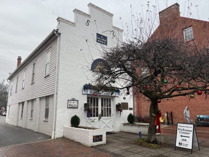 A small white building sits alongside a red brick walkway with a short tree directly in front of it. Slightly obscured by the tree branches, a sign above the doorway reads "Shepherdstown Welcome Center." There is a historic plaque to the side of the door with its text obscured by the distance.