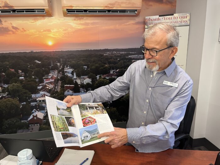 A man in a collared shirt and glasses stands behind a desk, holding open a pamphlet and pointing to one section of it.