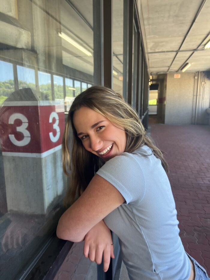 A young woman stands on a brick sidewalk leaning against a window. She smiles.