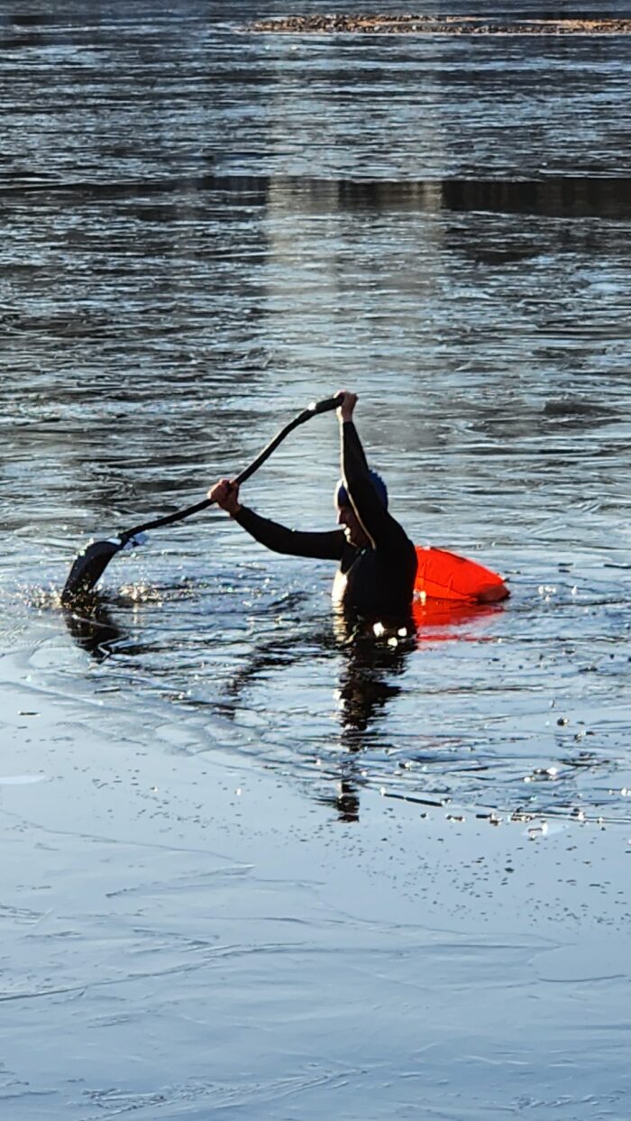 A man in a black wet suit is seen in water holding a shovel to break ice.