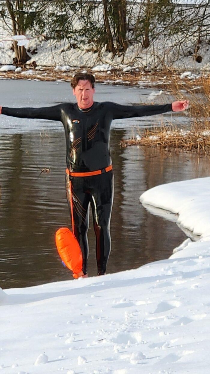 A man wearing a black wet suit stands on the shore, surrounded by snow, with his arms stretched out to the side.