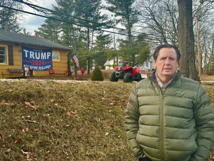 A man in an olive colored winter coat stands in front of a building with a large sign that reads, "Trump 2024 No More Bulls***."