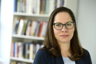 A photo of a women with dark brown hair posing for a photo. Behind her is a bookshelf that is blurred. She wears glasses.