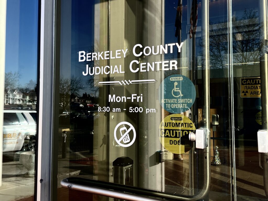 A glass door displays white text that reads "Berkeley County Judicial Center, Monday through Friday, 8:30 a.m. to 5 p.m." A parking lot is reflected in the glass door.