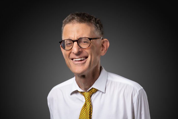 An older man with gray hair smiles for the camera in a posed photo. He wears a white dress shirt with a yellow tie and glasses.