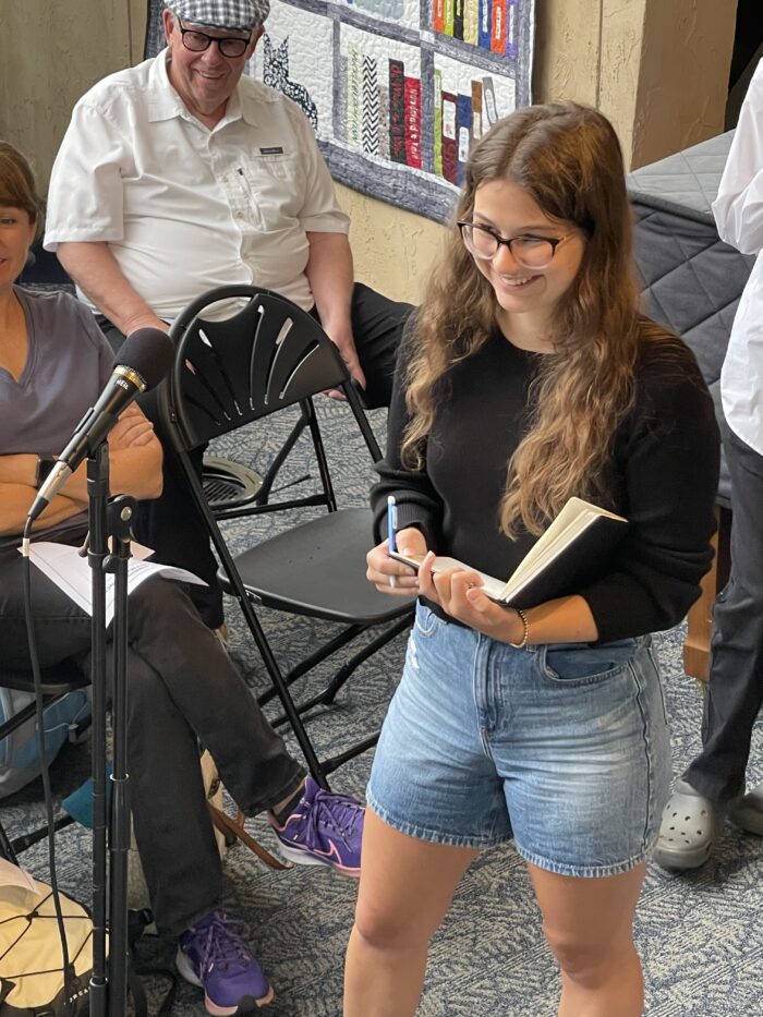 A young woman, smiling, with long brown hair, wearing shorts and a long-sleeved shirt, stands at a microphone in a room full of people. She wears glasses and holds a journal and a pencil.