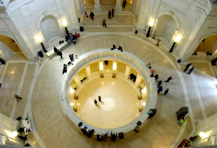 Viewed from above, people in formalwear stand and walk about a white marble rotunda. It is two stories, with a bottom floor visible through a circular hole in the middle of the room, overlooked by a marble wall along its perimeter upstairs.