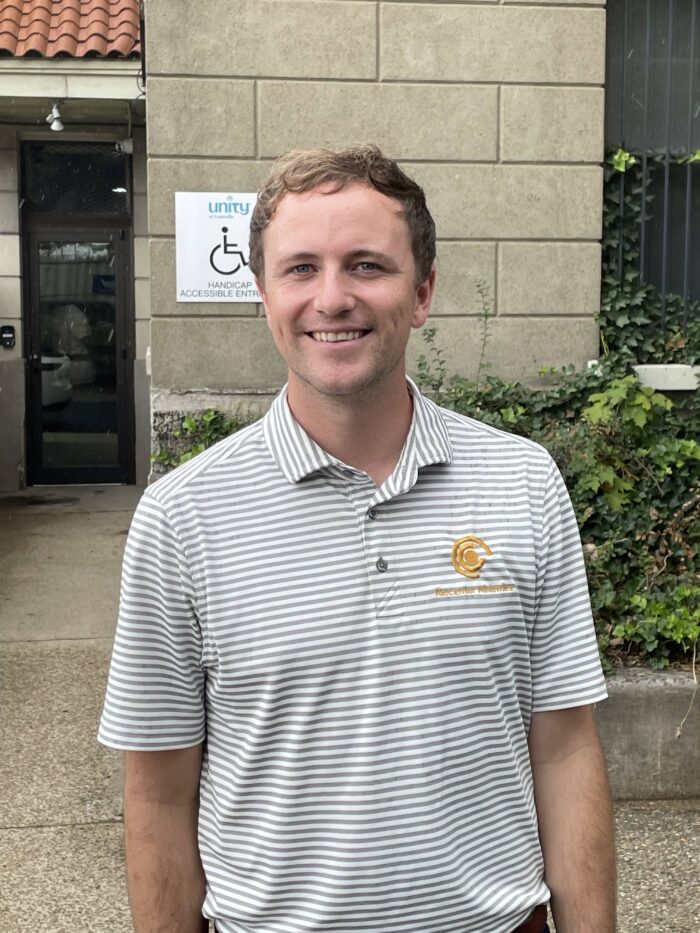 A young adult man smiles for the camera. He has short brown hair and wears a white shirt with gray stripes.