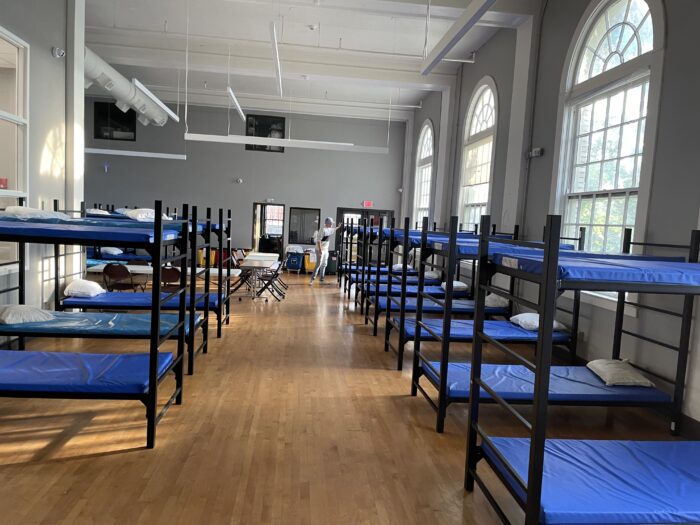 Rows of empty beds with blue mattresses are seen in a room with high ceilings and large windows.