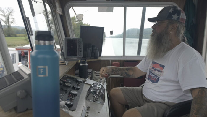 A man with a salt and pepper beard, tattoos, and ball cap with the American flag on it, steers a ferry. The man has his ears pierced and is wearing glasses.