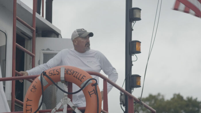 A man with a trimmed beard and mustache stands on the deck of a ferry. He looks out, away from the camera. The sky is overcast, and he wears a white long-sleeved shirt and ball cap.