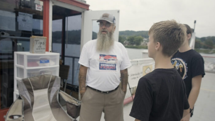 A man with a long, salt and pepper colored beard stands on the deck of a ferry with his hands in his pocket. Two young people, a boy and a girl, stand near him.