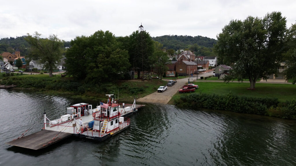 A red, white, and black colored ferry is seen next to dock. An American flag is flying above it. On shore, there is a neighborhood and trucks parked in grass. The sky is overcast.