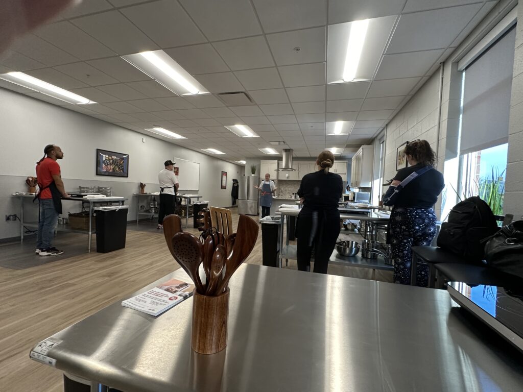 A container of kitchen utensils stands in the back of a teaching kitchen as students watch an instructor demonstrating proper cooking techniques.