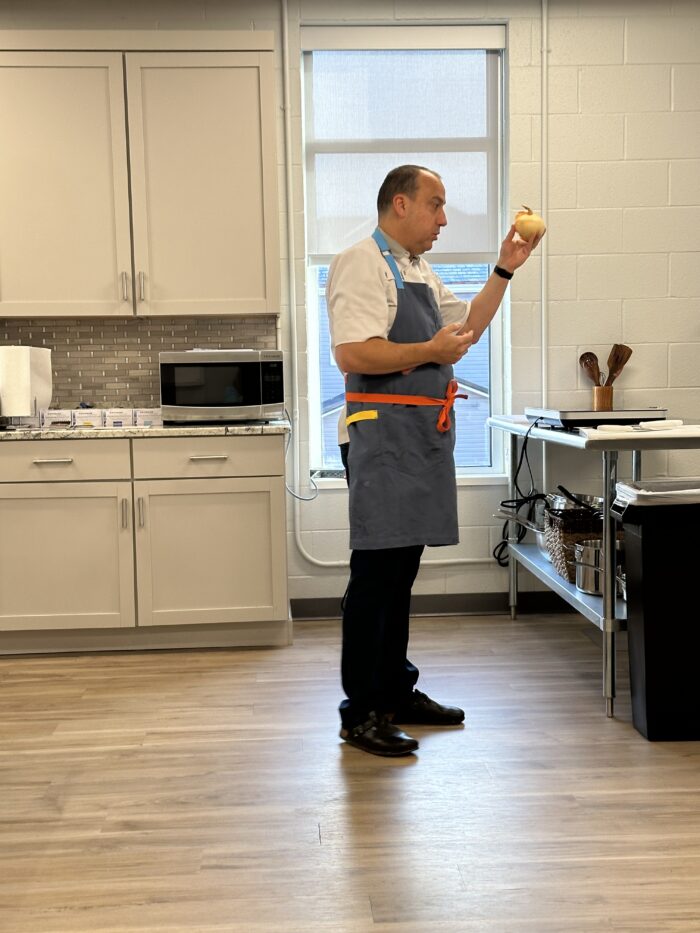 A man wearing an apron stands at the front of a teaching kitchen holding an onion.