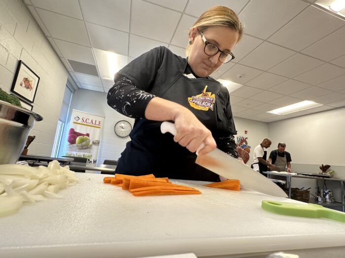 A woman wearing a black shirt, glasses and a ponytail uses a large kitchen knife to slice carrots and celery. Maria Young/West Virginia Public Broadcasting