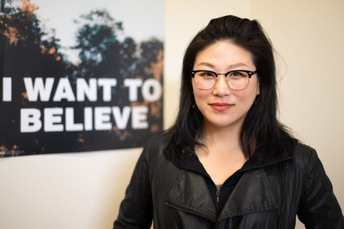 A young adult woman with dark black hair and glasses poses for a photo. A poster is seen behind her that reads, "I want to believe."