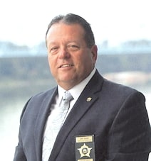 A headshot of a man with gray hair wearing a dark blazer, tie, and white dress shirt. He has a sheriff's badge on his chest.